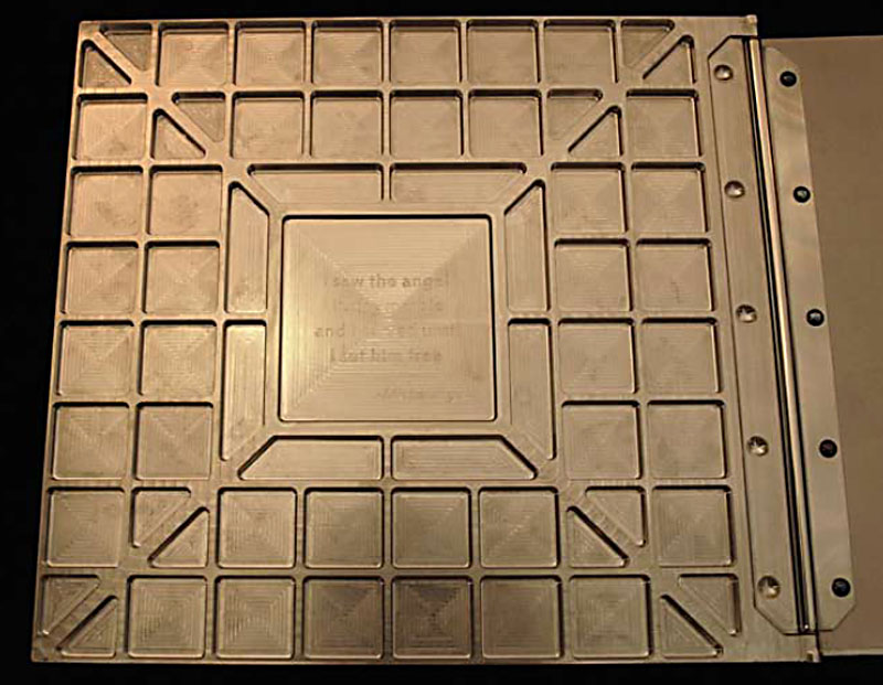 A square metal plate with a grid etched in metal featuring an inscription: "How the angel and the devil end up on his side".