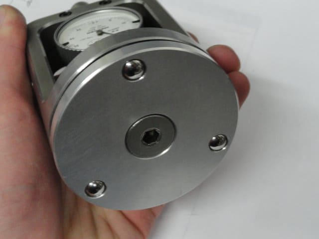 A silver bearing base with four evenly spaced holes and a white dial gauge displaying inch and millimeter measurements.