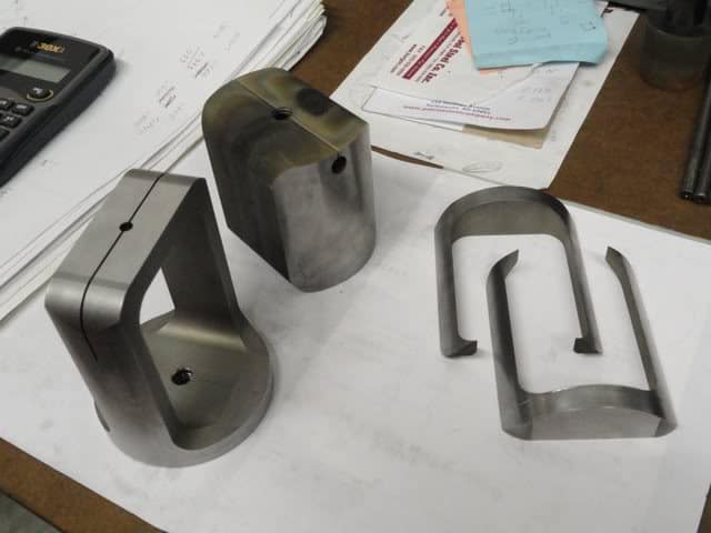 Three dark gray metal parts on a white workbench or table in a machine shop.