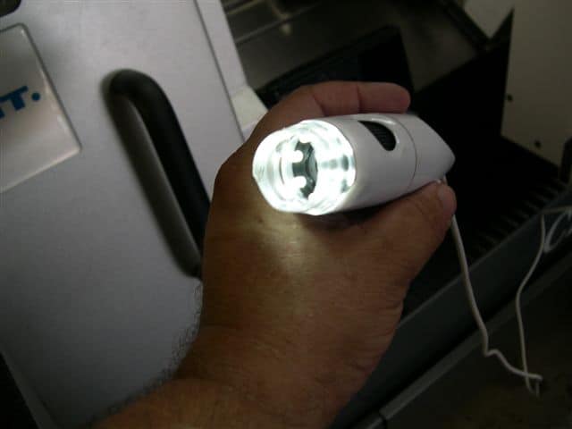 A hand holds a compact white digital microscope with a bright light illuminating its subject.