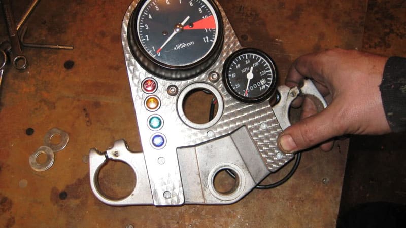 Motorcycle speedometer and tachometer gauges on a metal plate with a hand holding an object.