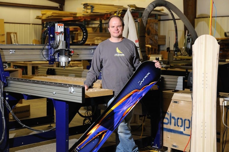 A man in casual attire proudly holds up a snowboard in a workshop with a large metal machine in the background.
