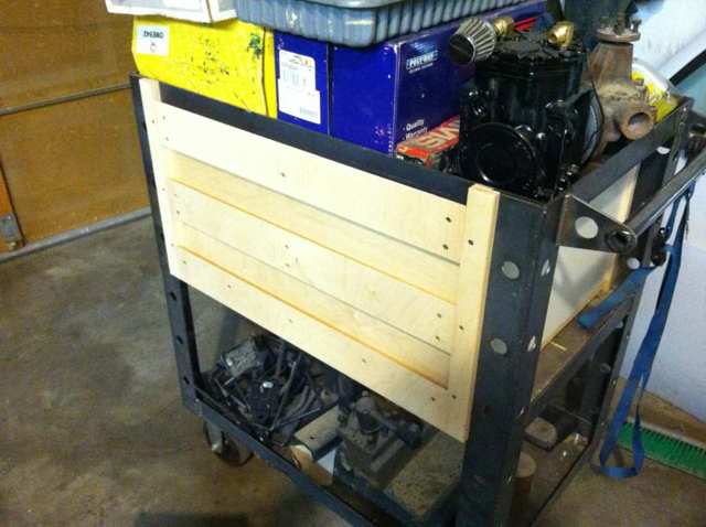 Metal cart with wheels in front of an open garage door, yellow box on top, white board attached to side secured by bolts.
