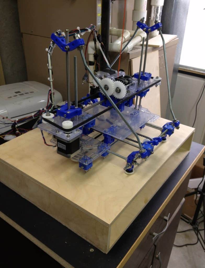 A 3D printer on a wooden platform with blue components and clear acrylic parts, set against a backdrop of workshop equipment.