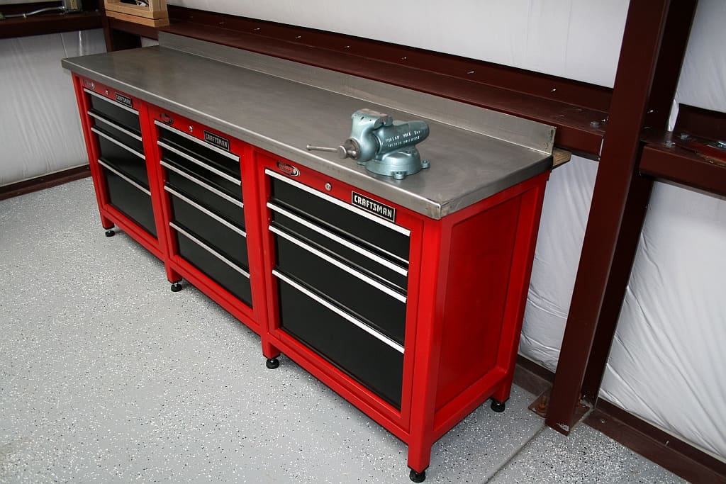 Red metal workbench with black drawers and stainless steel top, featuring "Craftsman" logo. Equipped with wheels and blue vice on white wall.