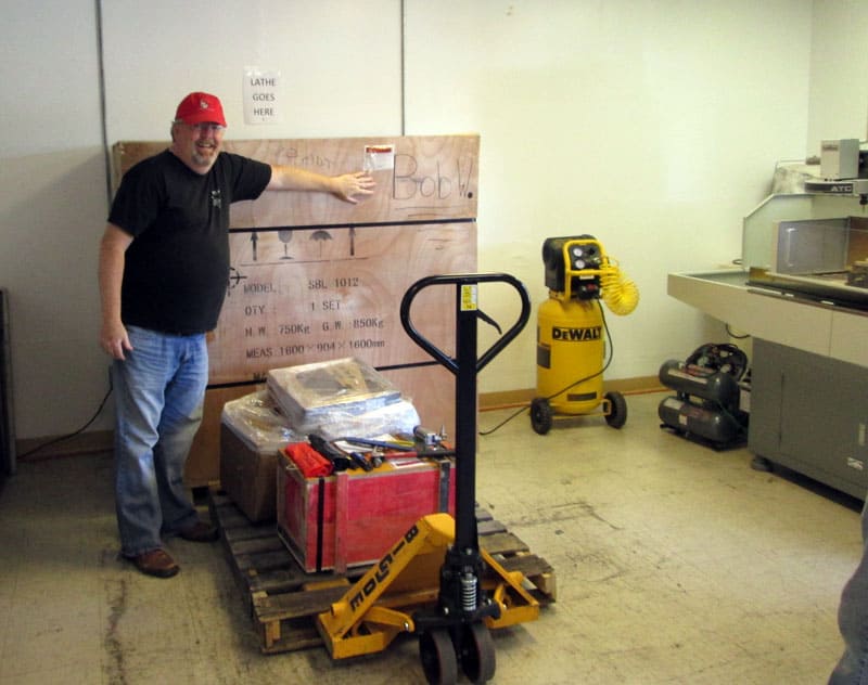 A man in a red hat stands in front of a wooden crate with black text, next to an orange pallet jack and a yellow air compressor in a workshop setting.