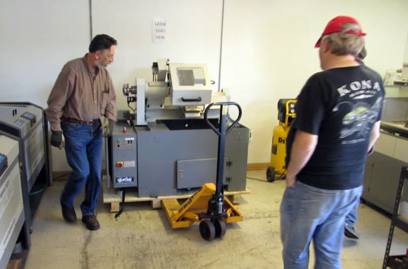 Man in blue jeans and brown shirt standing beside a large metalworking machine in a factory or workshop setting.
