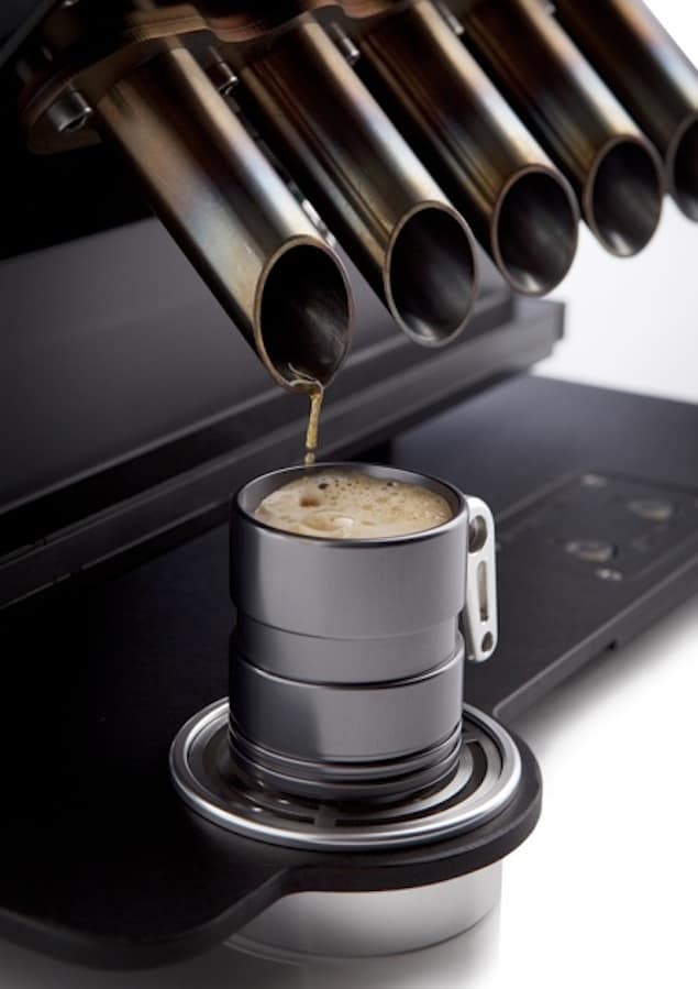 A stainless steel cup sits on top of a metal device with brown liquid pouring in, set against a dark gray background.