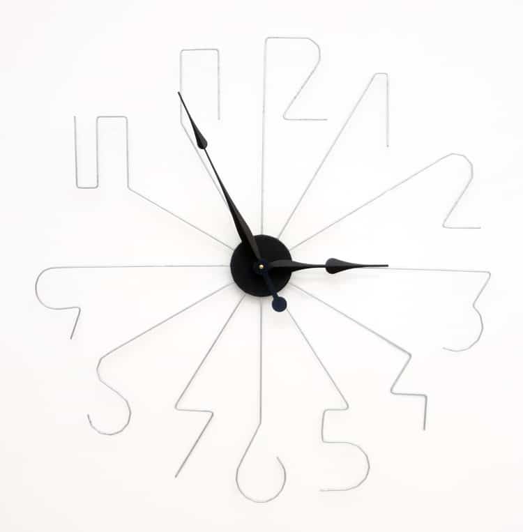 A black clock with thin wire hands and numbers, resembling a spider's web, against a plain white background.