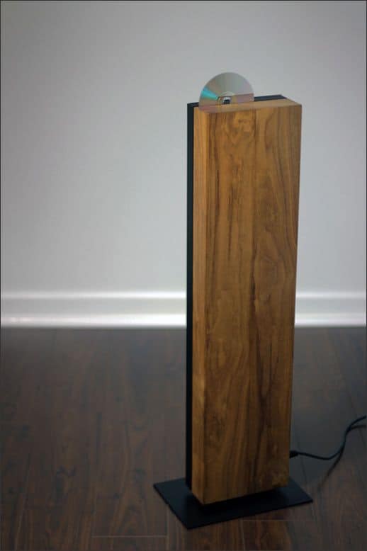A wooden speaker stand with a rectangular base and tall column against a white wall, featuring a light brown wood finish and black metal plate.