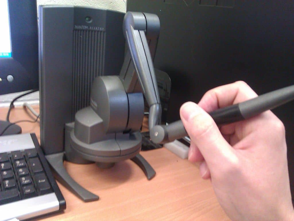 A hand grasping a black haptic arm in front of an office computer setup.