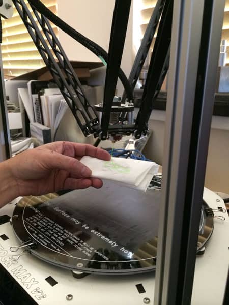 A close-up photograph of a 3D printer's extruder head with nozzle and surrounding components on a white platform.