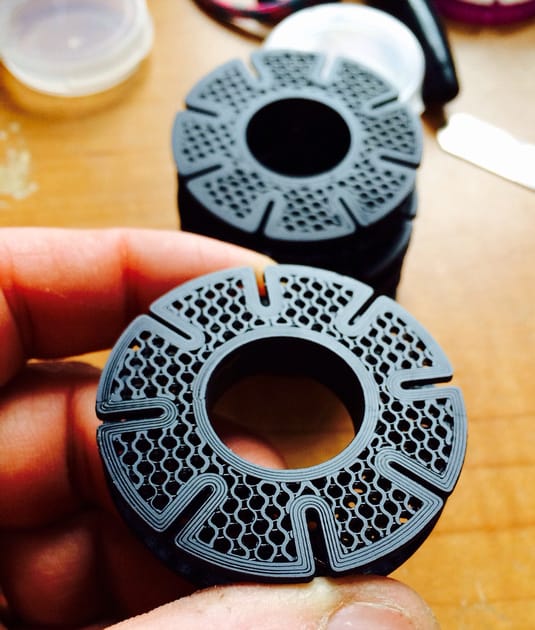 A close-up photograph of a small, black, circular part with a hole and honeycomb pattern on its surface.