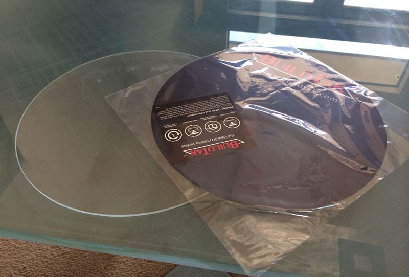 A vinyl record on a glass table with a black BuildTak sticker and plastic wrap, featuring white text "The next 3D printing surface".