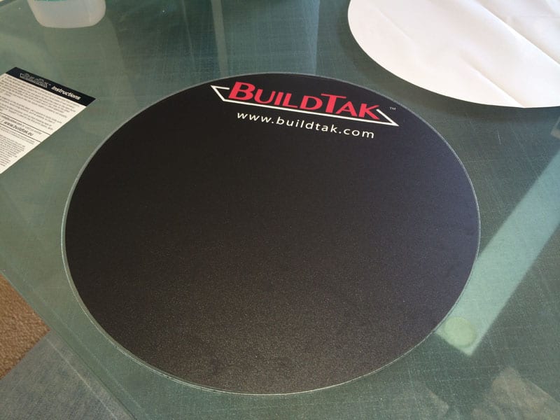 A circular BUILDTAK mat with red text and white logo design on a glass surface.