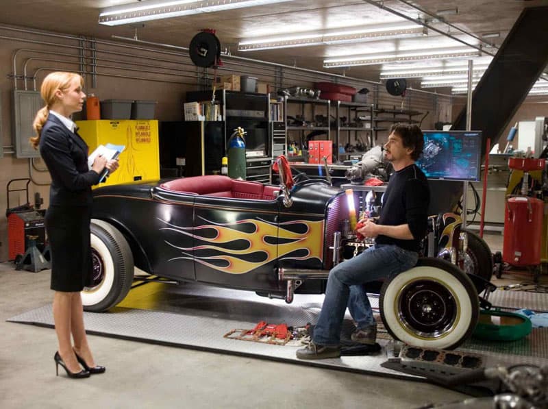 A couple standing next to a black customized car with a man sitting on it in a garage workshop setting.