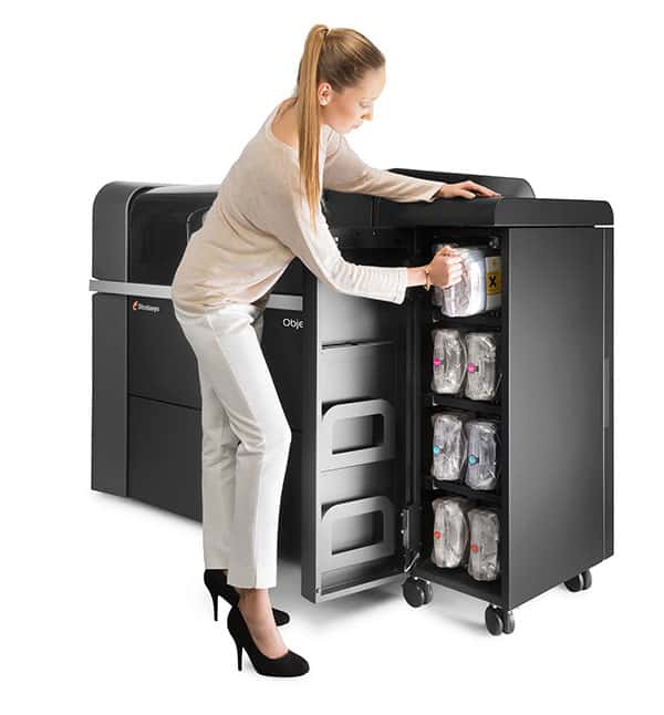 A woman in business attire stands beside a large black 3D printer with an open door showing plastic bags.