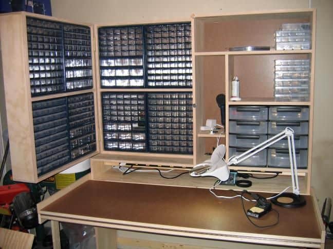 Well-organized workspace with wooden table and shelving units containing drawers and containers for metal fabrication supplies.