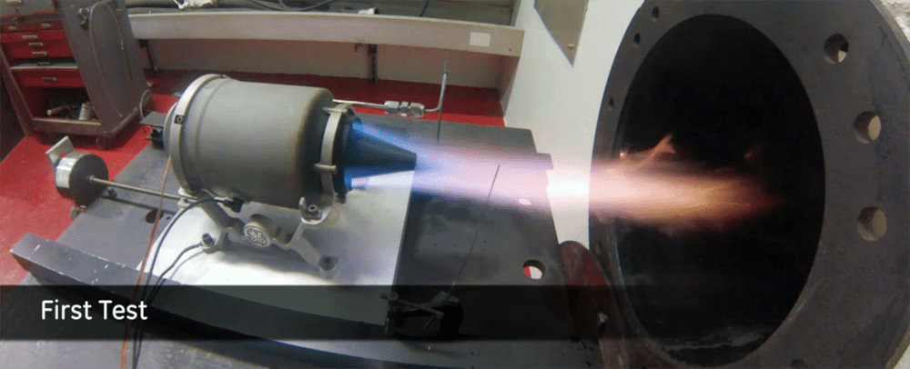 A 3D printed mini jet engine on a metal base plate with flames emanating from its nozzle.