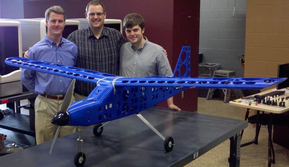 Four men standing behind a large blue drone with propellers in an office or workshop setting.