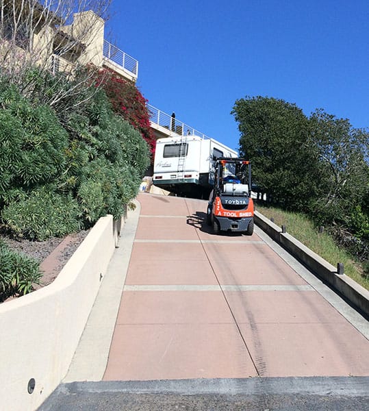 Forklift on concrete ramp with bushes, trees, and a white camper against a blue sky.