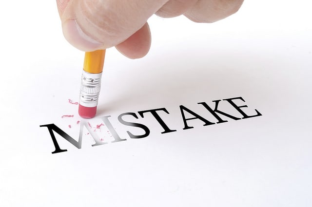 A hand rubs out the word "MISTAKE" with a yellow pencil and red eraser.