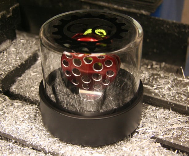 A glass cylinder with a black base and a red object with circular holes sits on a metal surface covered in shavings.