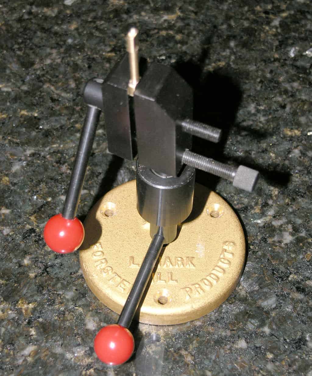 Small metal vise with red balls on legs, brass-colored base plate and granite background.