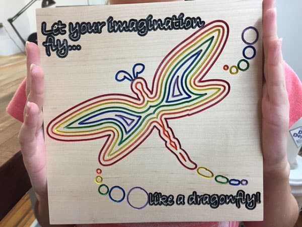 A wooden sign with a colorful dragonfly design and inspirational text, featuring a stylized dragonfly in pastel colors.