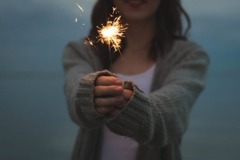 Woman holding lit sparkler with flame casting warm glow on her face.