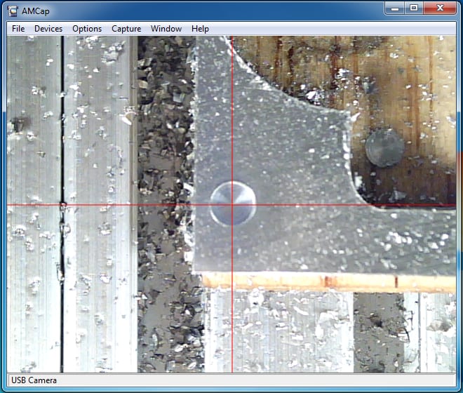 A red crosshair overlay in an AMCap window on a wooden workbench with a metal object.