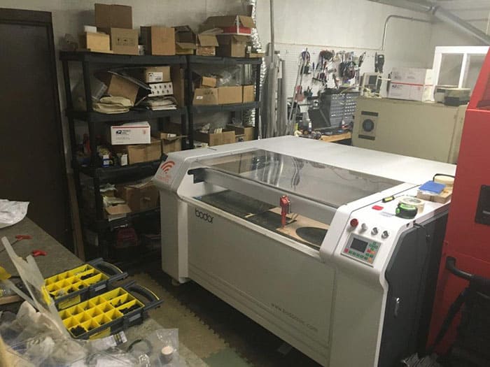 A cluttered workshop with a large white laser cutter, shelves stacked with boxes and bins, and a floor covered in cords, tools, and materials.