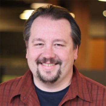 Dark-haired man with mustache in red plaid shirt and black t-shirt.