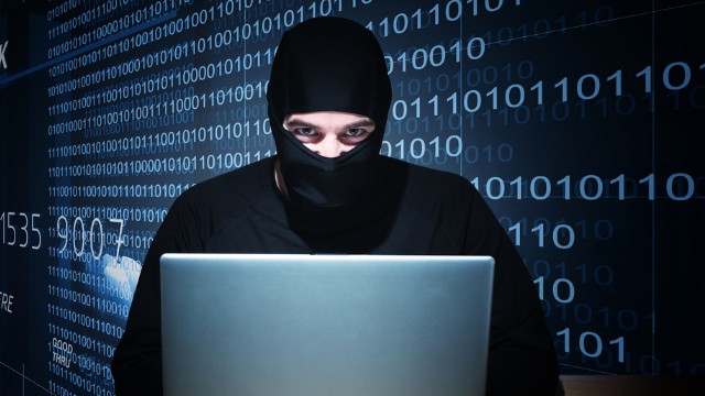 Person wearing black balaclava seated in front of illuminated computer screen with binary code background.