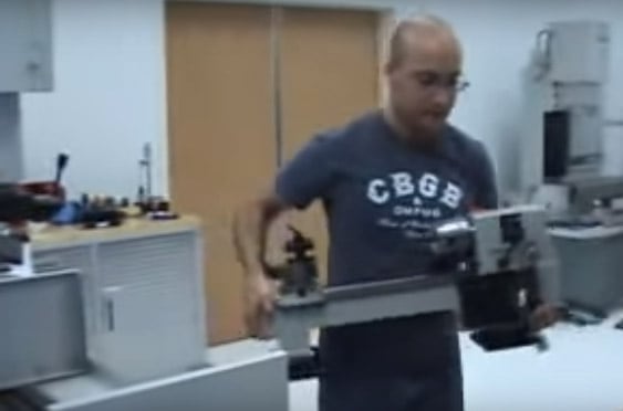 A bald man wearing glasses and a dark blue t-shirt with white letters, holding a lathe in a workshop.
