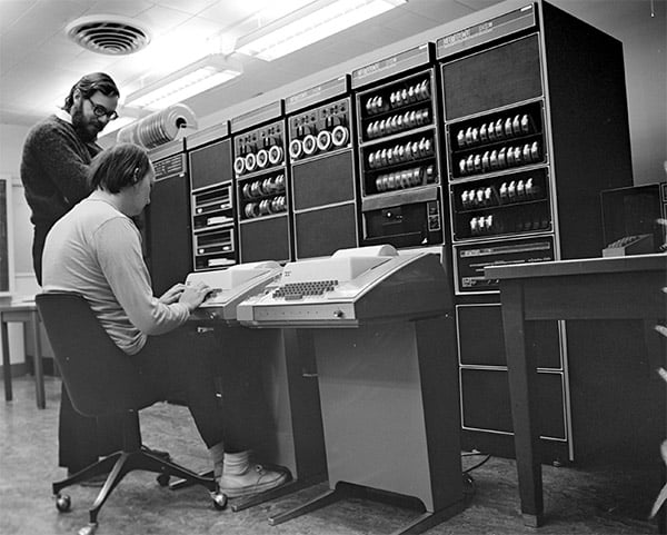 Two men working on an old computer system with fluorescent lighting.