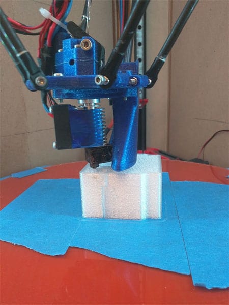 A 3D printer bearing block being printed on a blue platform with red and black wires in the background.