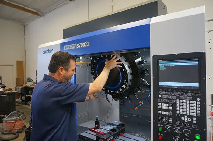 A man operates a Brother S700XV Speedio machine in a workshop/warehouse environment.