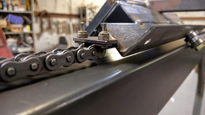 Dark gray metal chain with rusted links attached to bolts on a black metal frame in a cluttered workshop.