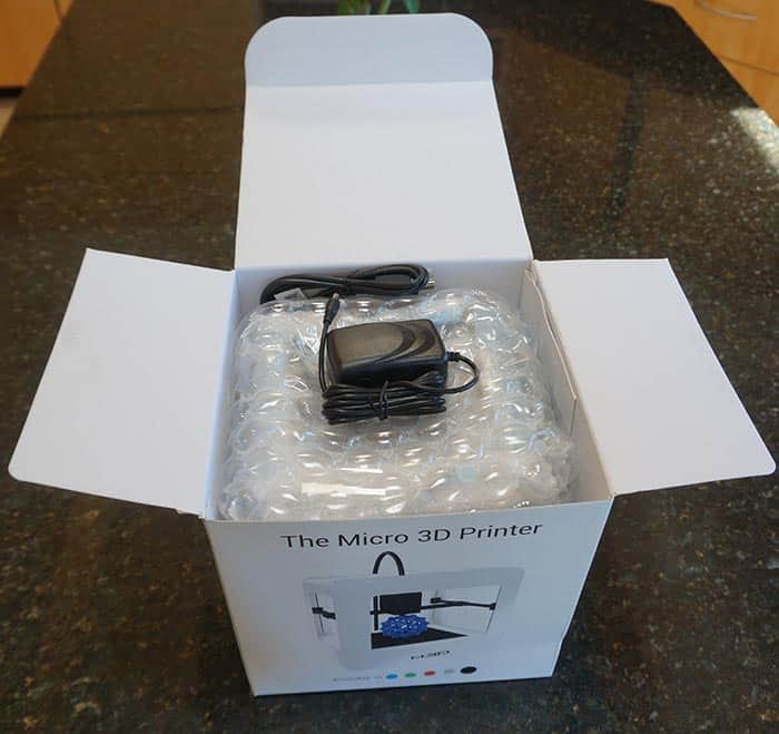 A white box with "The Micro 3D Printer" label and logo, containing a black device wrapped in bubble wrap and a power cord.