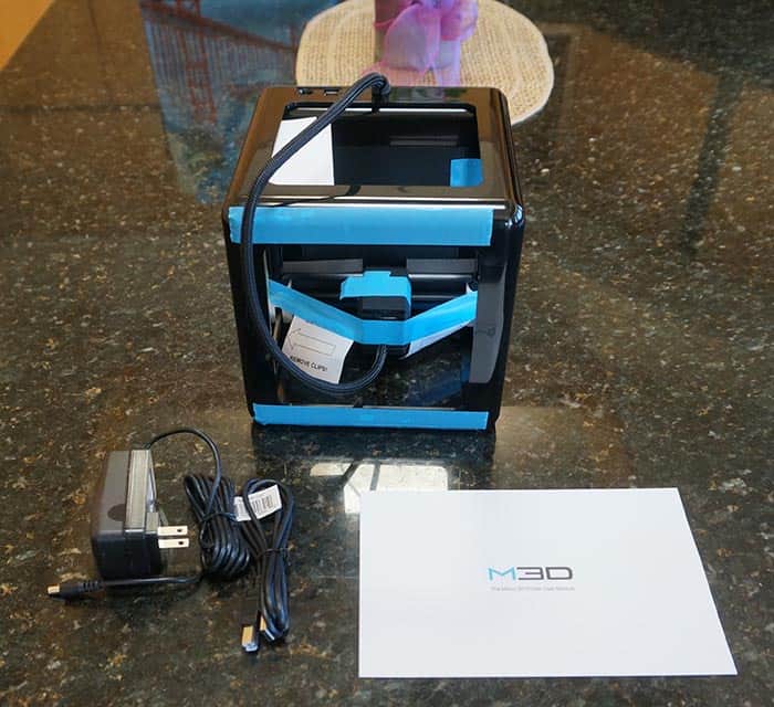 Black and blue plastic device with cords and wires on a dark surface, beside white paper with "M30" written on it.