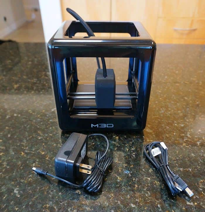 Black 3D printer (Monoprice Maker Select V2) with power adapter and USB cable on speckled countertop.