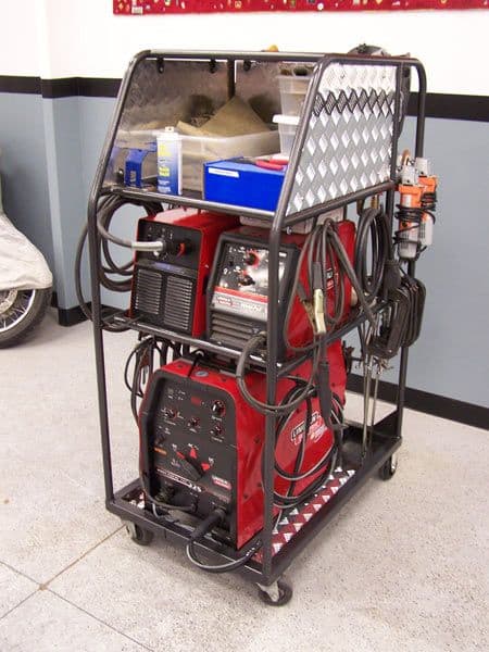 Red Lincoln Electric welder on mobile cart with tools and equipment in front of white wall with blue trim.
