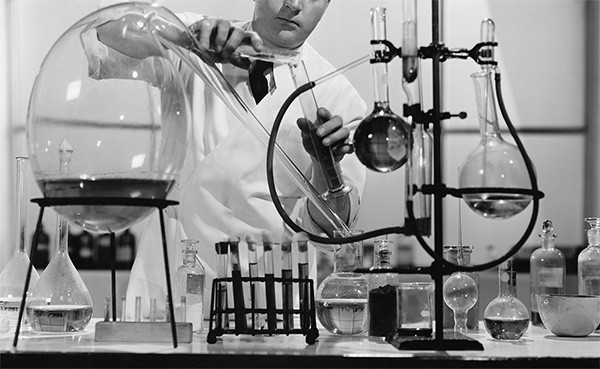 A man in a lab coat pours liquid from a beaker onto a cluttered laboratory counter.