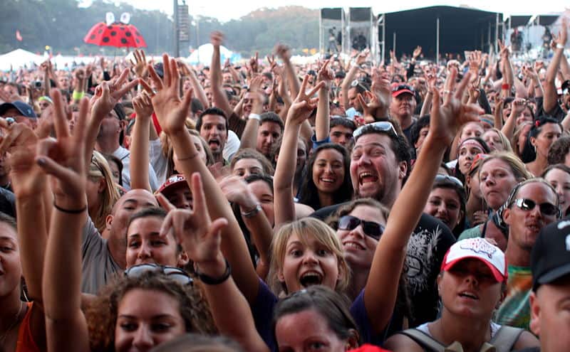 Densely packed crowd at an outdoor festival with ladybug umbrella and distant tents.