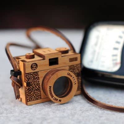 Wooden camera pendant with compact design, light-colored wood body, and intricate carvings.