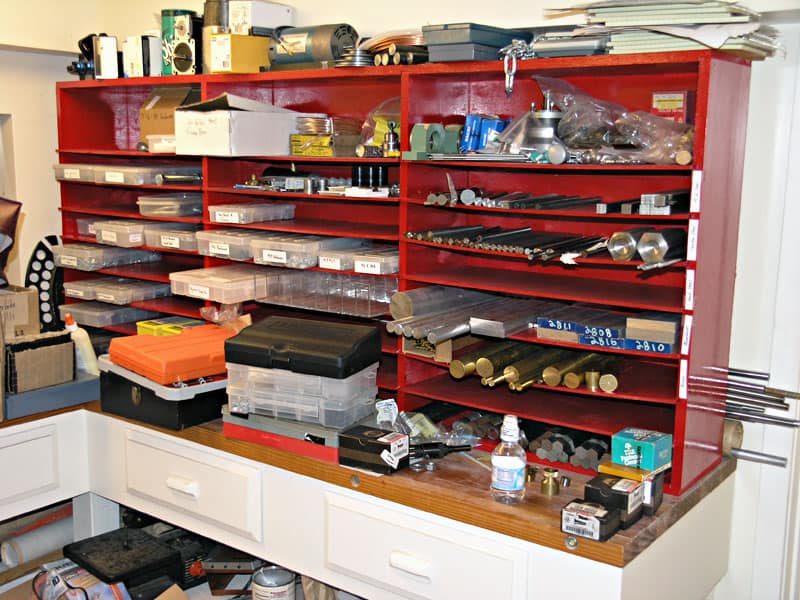 Cluttered workshop storage room with red shelving unit holding various tools and machinery parts.