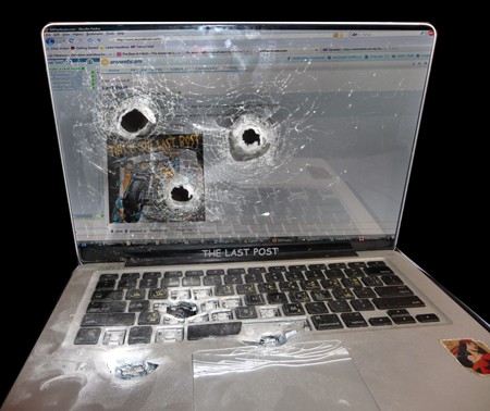 Damaged laptop with bullet holes in screen and cracked keyboard against gray background.