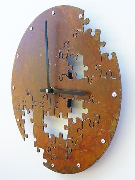 Rustic puzzle clock with irregularly shaped pieces cut out to form a clock face on a white wall.
