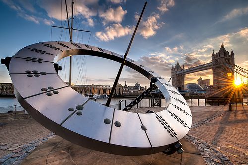 Sundial sculpture near Tower Bridge, London, with cloudy sky and Roman numeral markings.